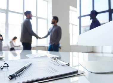 A signed contract sits on a desk and two men shakes hands in the background.