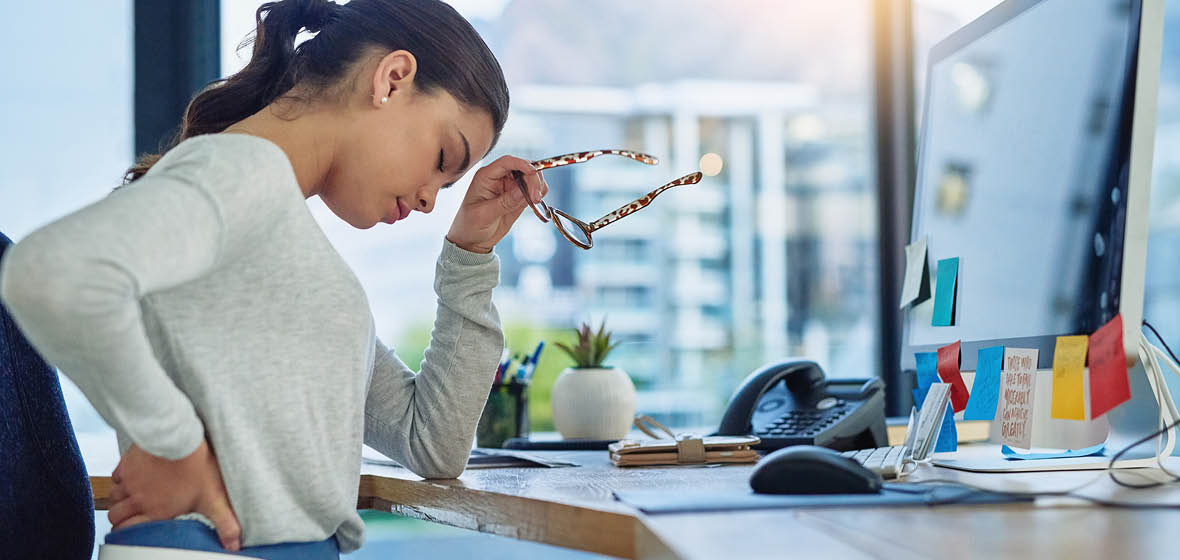 woman in pain holds her back, sitting at a desk in an office