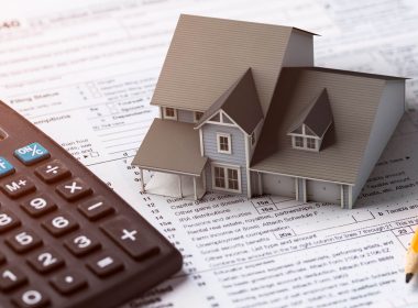 Scale model of a house sits on top of a a checklist on a desk, next to a pencil and a calculator.