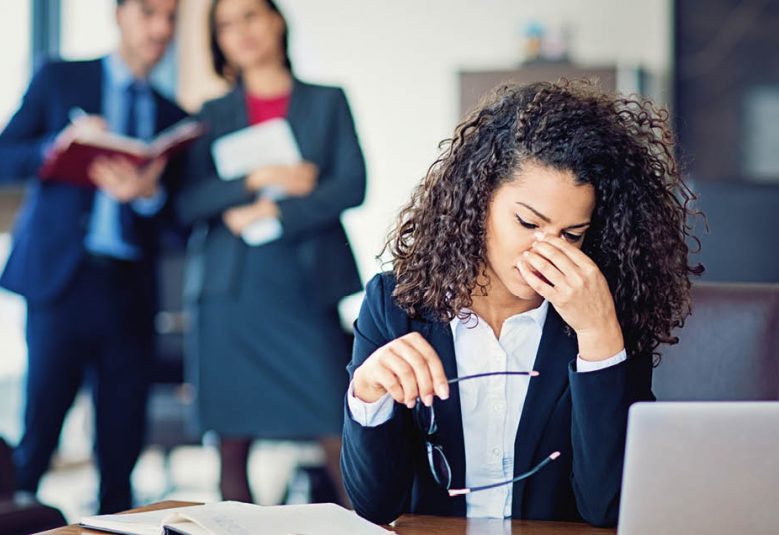 Stressed woman at desk with colleagues looking at her from a distance