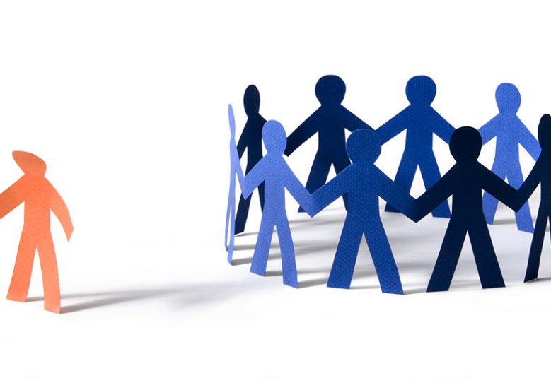 Orange paper person with dejected stance off to the side of a circle of blue paper people.