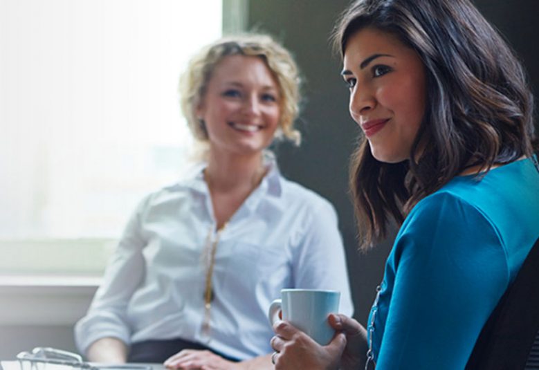 two women seated at a desk talking over coffee