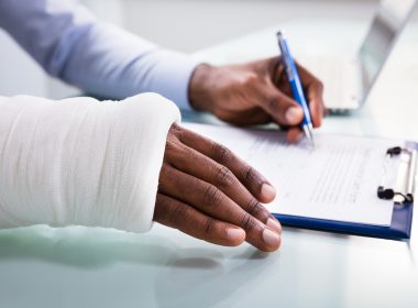 Overhead View Of Injured Man With Bandage Hand Filling Insurance Claim Form On Clipboard
