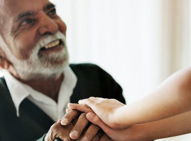 Elderly man sits on a chair with a young person holding his hand