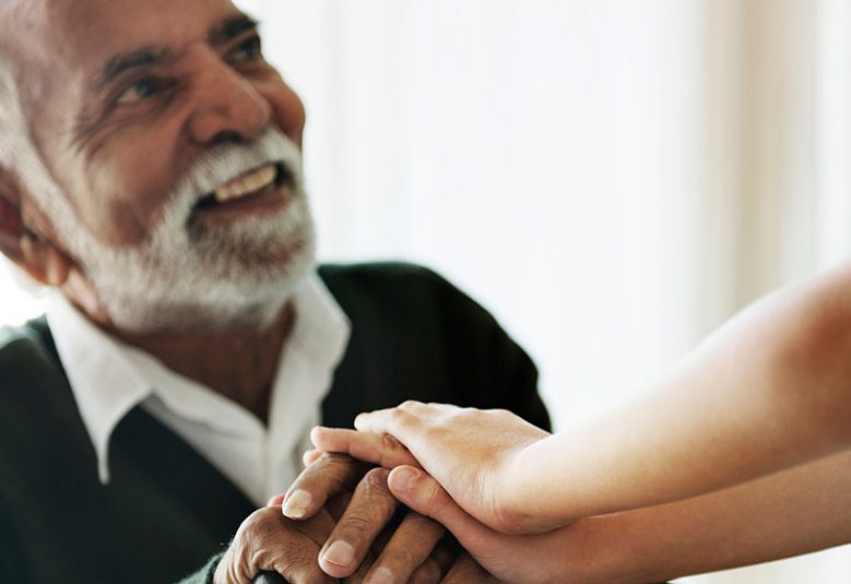 Elderly man sits on a chair with a young person holding his hand